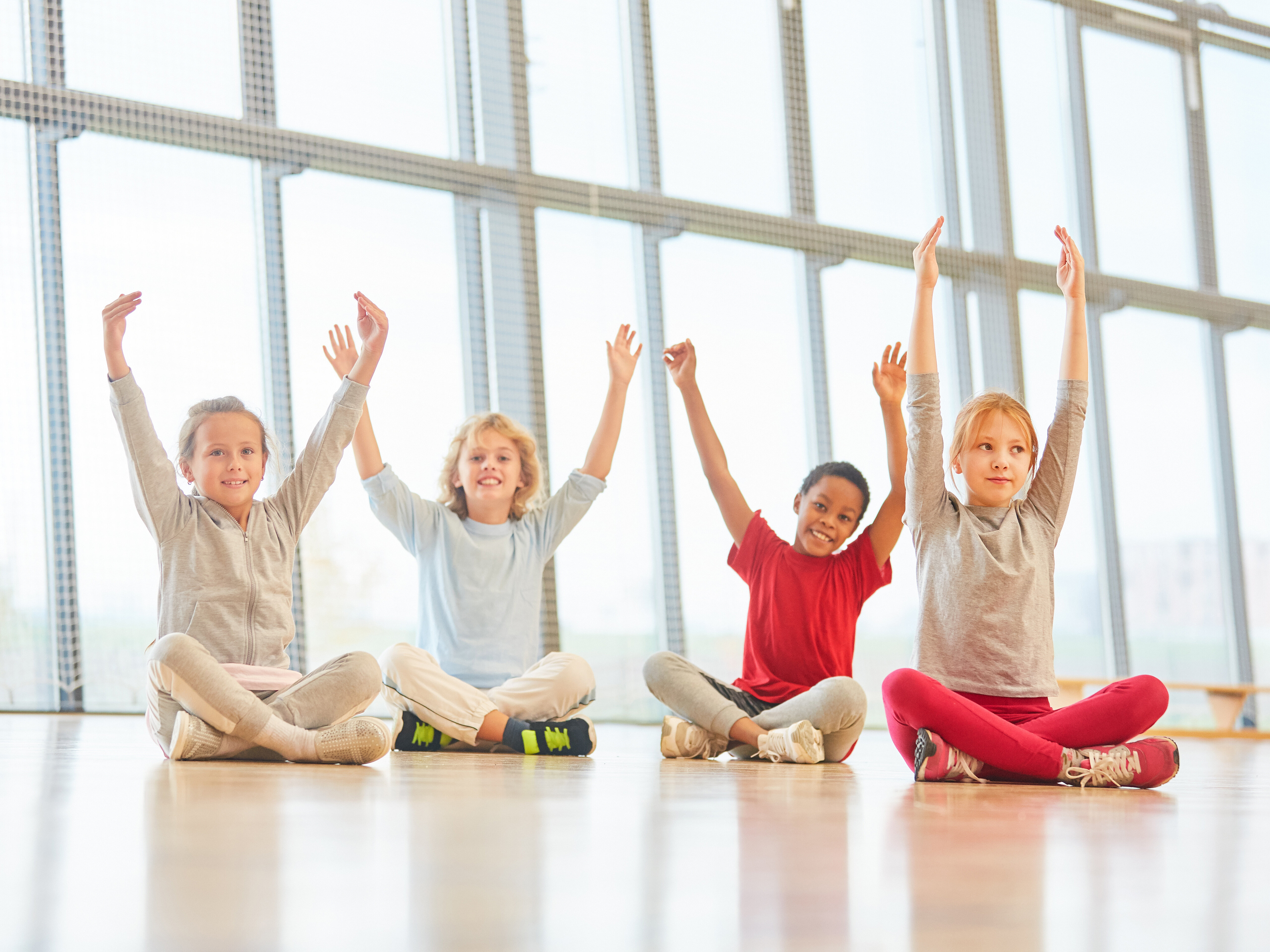 Vier Kinder sitzen auf dem Boden in einem hellen Raum mit großen Fenstern. Sie haben die Arme erhoben und lächeln. Die Kinder tragen bequeme Kleidung in verschiedenen Farben und scheinen Freude zu haben.