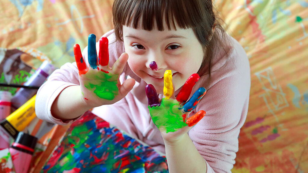 Ein Kind mit Down-Syndrom zeigt stolz seine bemalten Hände, die mit bunten Farben in verschiedenen Tönen bedeckt sind. Im Hintergrund sind Malutensilien und ein farbenfrohes, gemustertes Bettlaken zu sehen. Das Kind lächelt und wirkt glücklich.