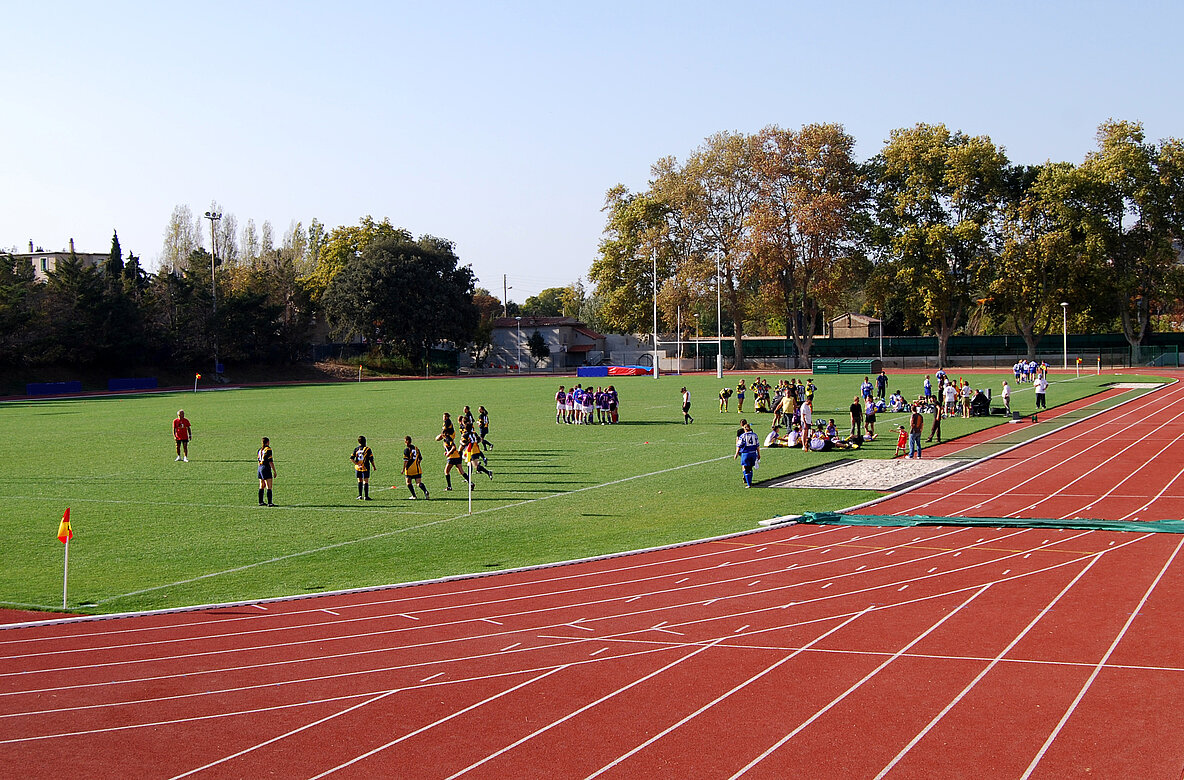 Eine Sportanlage mit einem Fußballfeld und einer Laufbahn. Auf dem Feld sind mehrere Gruppen von Spielern in verschiedenen Trikots, die sich auf das Spiel vorbereiten. Im Hintergrund sind Bäume und Zuschauer zu sehen.