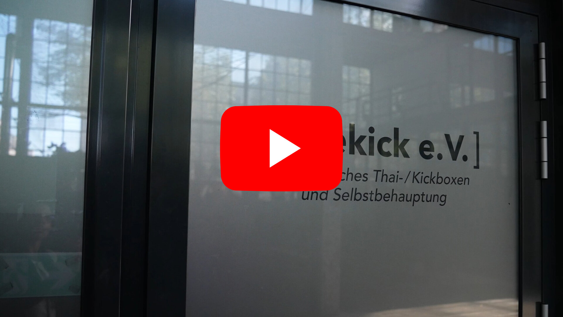 Eine Glaswand mit schwarzem Rahmen, auf der in schwarzer Schrift der Vereinsname 'Sidekick e.V.' sowie der Zusatz 'Deutsches Thai-/Kickboxen und Selbstbehauptung' zu lesen sind. Im Hintergrund sind unscharf Fenster und Gebäudestrukturen zu erkennen.
