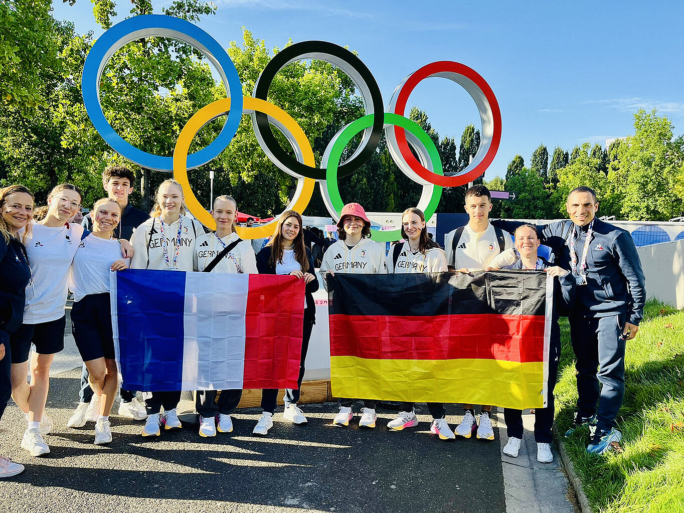 Eine Gruppe von Sportlern steht vor den Olympischen Ringen und hält die Flaggen von Frankreich und Deutschland. Sie lächeln und posieren für das Foto, umgeben von grüner Landschaft.