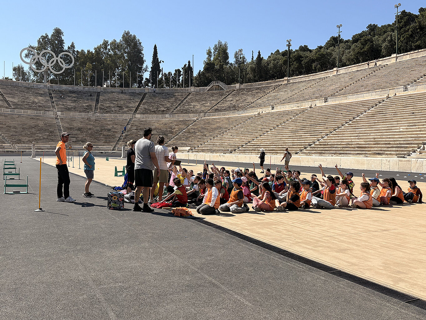 Eine Gruppe von Kindern in orangefarbenen T-Shirts sitzt auf dem Boden eines Stadions. Einige Erwachsene stehen vor ihnen und erklären etwas. Im Hintergrund sind die Olympischen Ringe und leere Sitzreihen zu sehen.