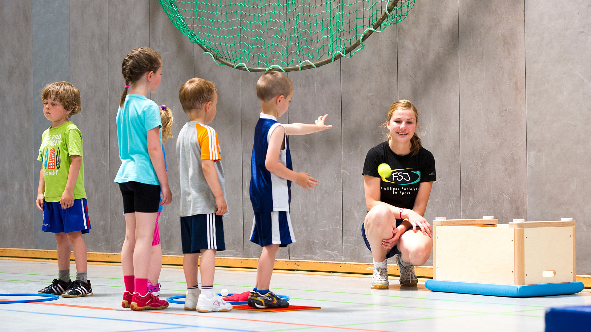 Eine Gruppe von fünf Kindern steht in einer Sporthalle, während eine Trainerin auf dem Boden sitzt und ihnen Anweisungen gibt. Die Kinder tragen sportliche Kleidung und scheinen aufmerksam zuzuhören. Im Hintergrund ist ein Netz sichtbar.
