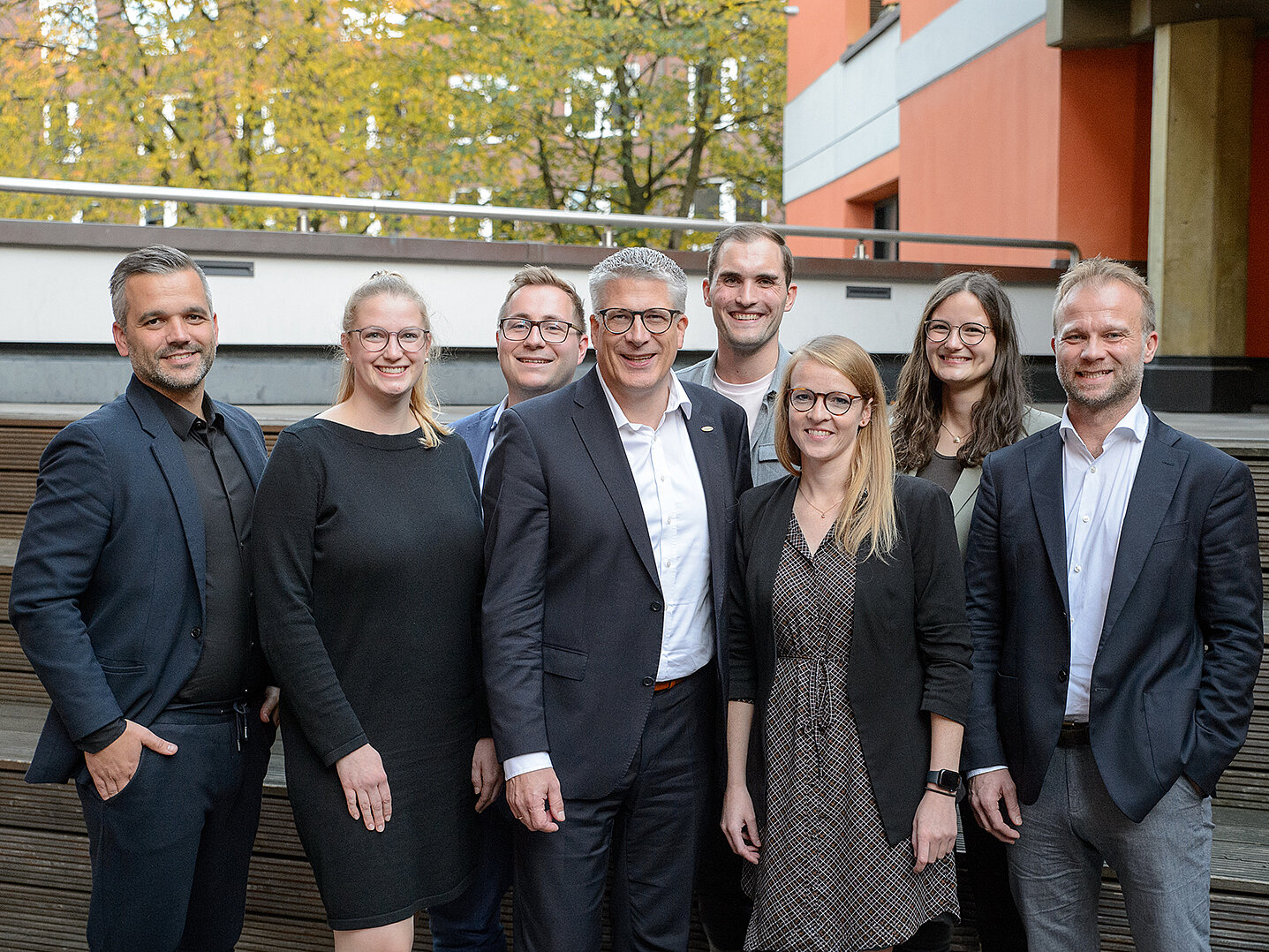 Eine Gruppe von acht Personen steht zusammen im Freien, umgeben von Bäumen und einem modernen Gebäude. Sie lächeln und tragen formelle Kleidung. Die Szene vermittelt eine freundliche und professionelle Atmosphäre. Laut Kontext zeigt das Bild den Vorstand der dsj.