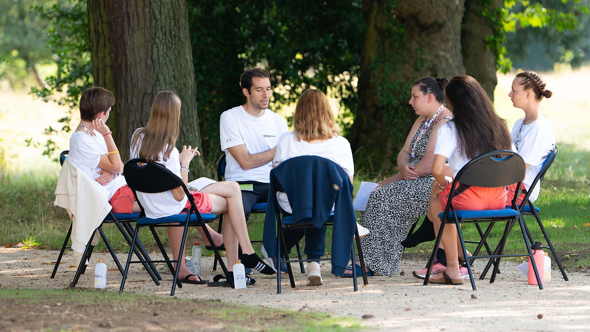 Eine Gruppe von acht jungen Personen sitzt im Freien auf Stühlen in einem Kreis. Sie diskutieren miteinander, während Bäume im Hintergrund sichtbar sind. Einige Teilnehmende tragen T-Shirts, und es sind Wasserflaschen auf dem Boden zu sehen.