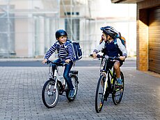 Zwei Kinder fahren auf Fahrrädern in einer gepflasterten Straße. Das erste Kind trägt einen Helm und hat einen Rucksack auf dem Rücken, während das zweite Kind ebenfalls einen Helm trägt. Beide lächeln und scheinen sich zu unterhalten. Im Hintergrund sind Gebäude und eine Garage sichtbar.