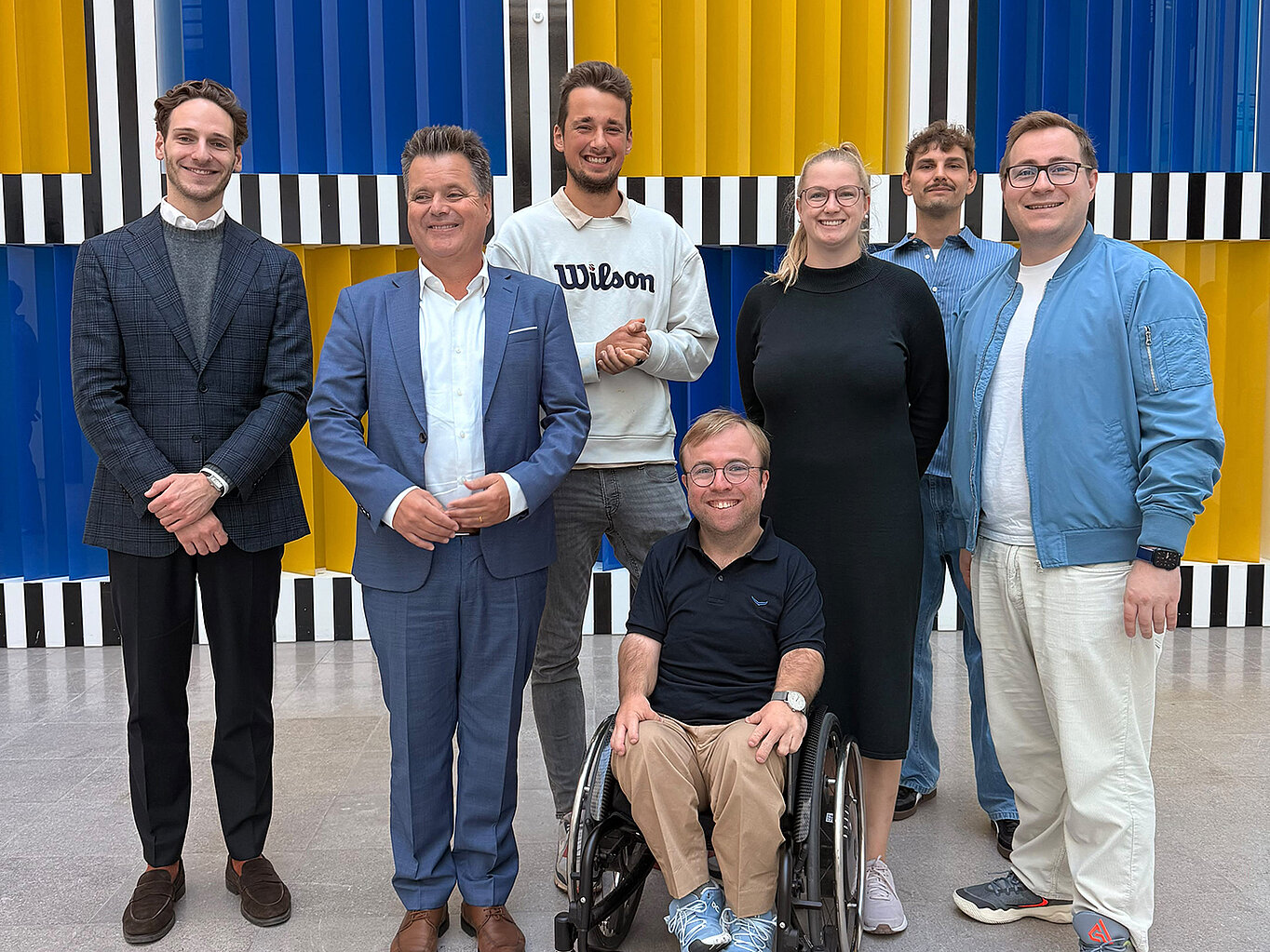 Eine Gruppe von sieben Personen steht vor einer bunten Wand mit gelben, blauen und schwarzen Streifen. Sie lächeln und posieren freundlich. Eine Person sitzt im Rollstuhl, während die anderen stehen. Die Atmosphäre wirkt freundlich und einladend.