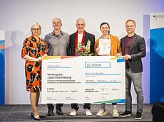 Eine Gruppe von fünf Personen steht auf einer Bühne und hält einen großen Scheck über 10.000 Euro. Der Scheck ist für den Vereinspreis "Sport mit Haltung" und wird an ESV Olympia Köln e.V. für ihre Leistungen im Sport verliehen.