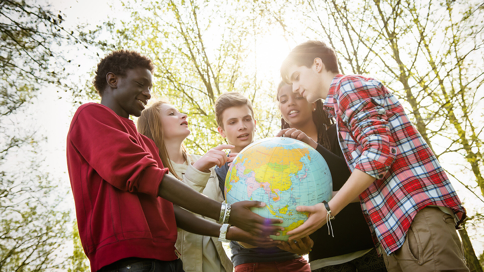 Sechs Jugendliche halten gemeinsam einen Globus, umgeben von Bäumen. Der Globus zeigt eine Weltkarte mit Kontinenten und Ländern. Fortbildung Sport kennt keine Grenzen für internationale Jugendbegegnungen im Sport.
