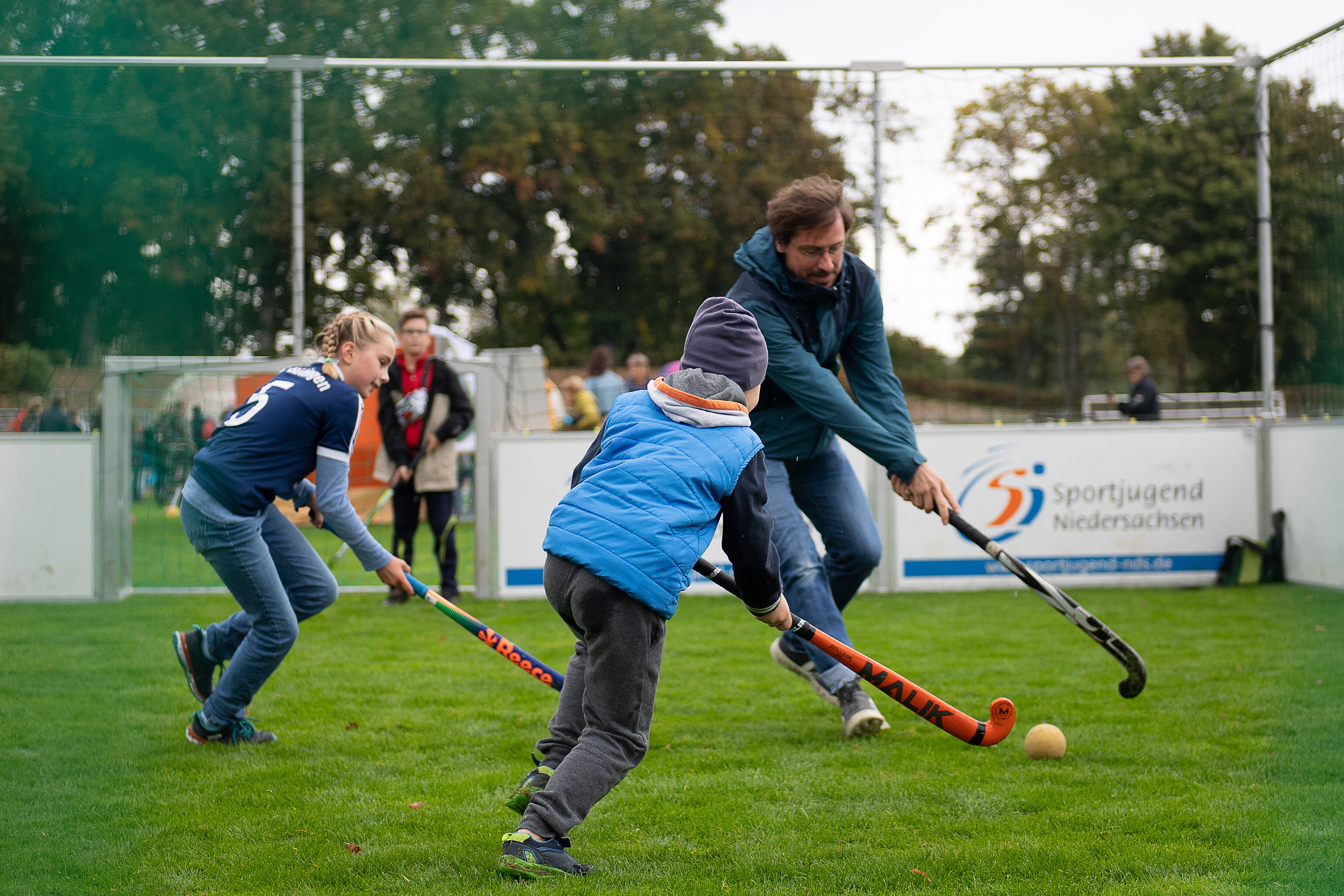 Drei Personen spielen auf einem Rasenfeld Hockey. Zwei Kinder, eines in blauer Jacke und eines in einem dunkelblauen Shirt, versuchen, den Ball mit ihren Schlägern zu treffen. Ein Erwachsener unterstützt sie dabei. Im Hintergrund sind weitere Personen und eine Werbetafel sichtbar.